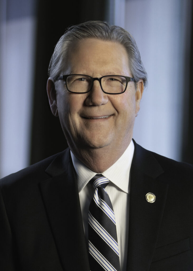 Gray-haired man with glasses, in a dark suit and striped tie, smiles indoors.
