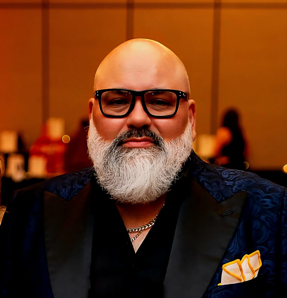 Bearded man with glasses in a suit and yellow pocket square poses indoors at an event.