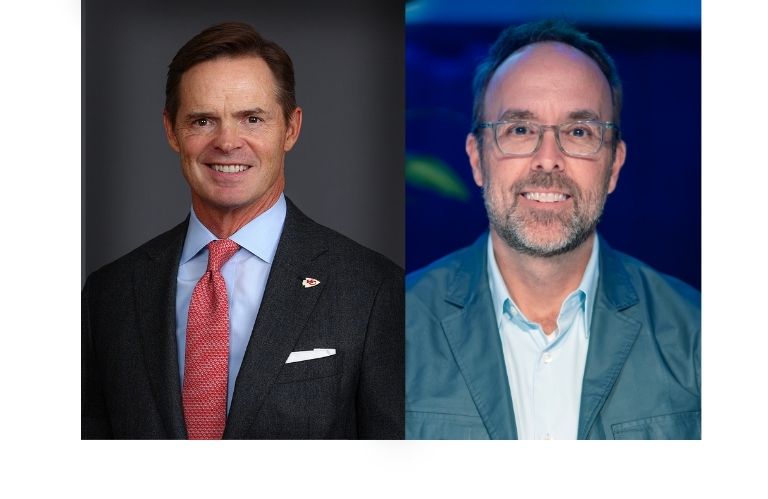 Two men pose: one in a suit and tie, the other wearing glasses and a casual jacket.