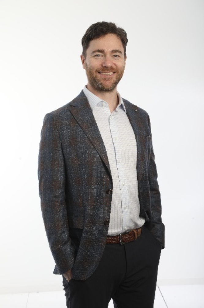 Smiling man with short brown hair and beard, in blazer and dress shirt, hands in pockets.