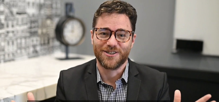 Smiling bearded man with glasses in a blazer sits indoors; clock visible behind him.