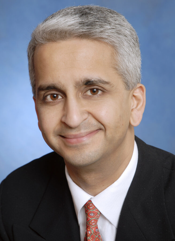 Smiling man with short gray hair in a suit and red tie, set against a blue background.