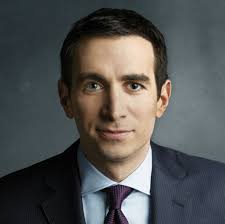 A man in a suit and tie with short dark hair stands against a simple dark background.
