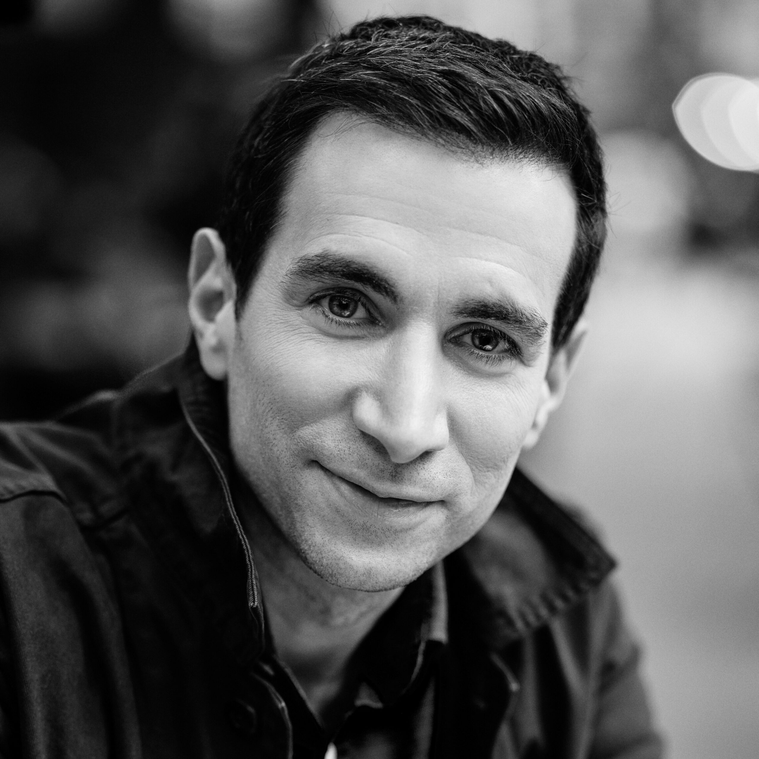 A man with short dark hair in a jacket smiles slightly in a black and white outdoor portrait.