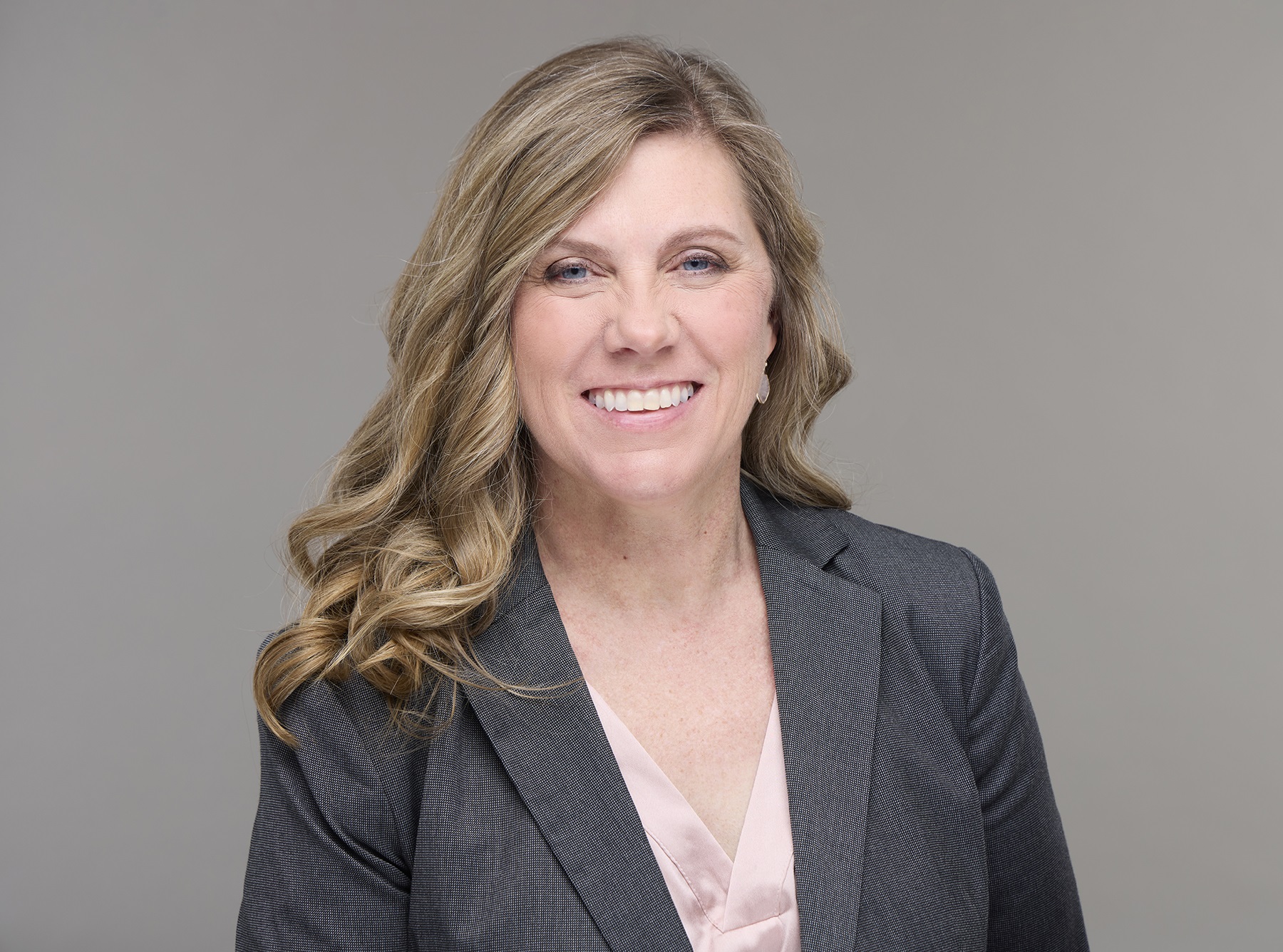 Smiling blonde woman in a grey blazer and blouse, standing before a plain background.