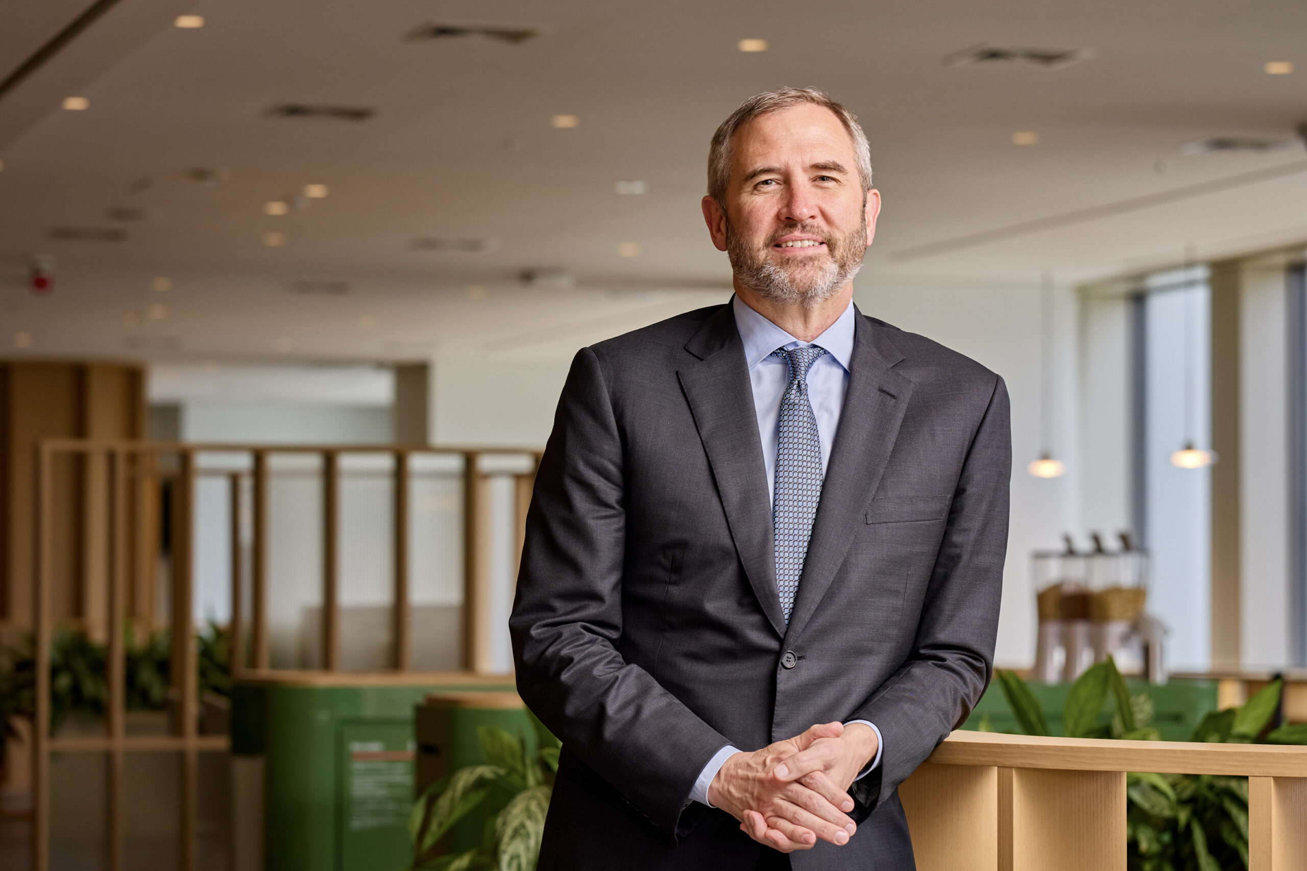 Smiling man in a suit stands indoors, surrounded by plants and modern office decor.