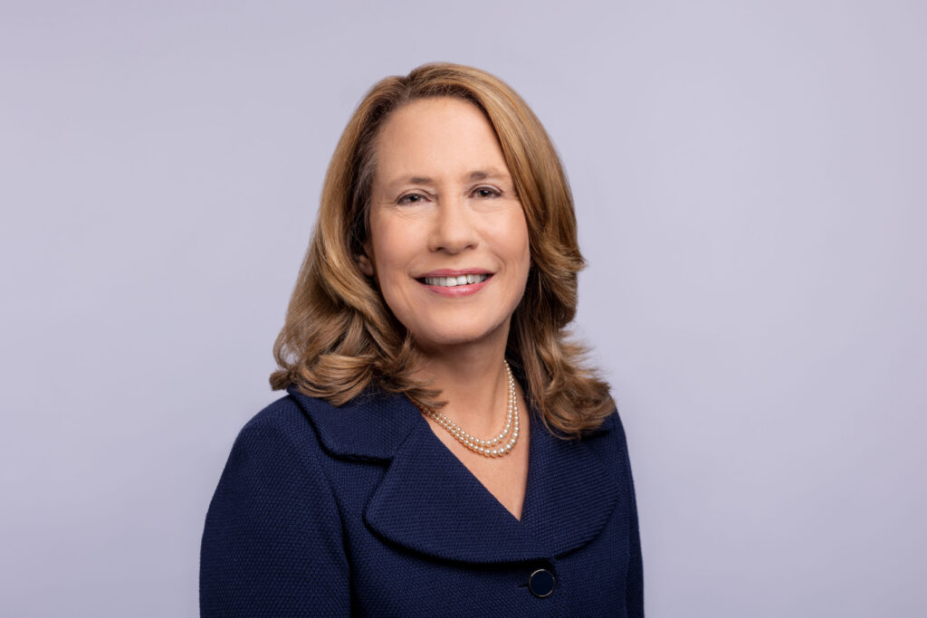 Smiling woman with shoulder-length light brown hair in a navy blazer, on a light gray background.